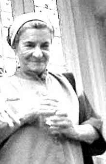 Fotografia em preto e branco de uma mulher branca, aparentando meia-idade, enquadrada da barriga para cima, com as mãos encostadas na altura do peito, trajando um vestido largo e usando uma faixa no cabelo. Ela está sorrindo. Ao fundo, vê-se uma parede.