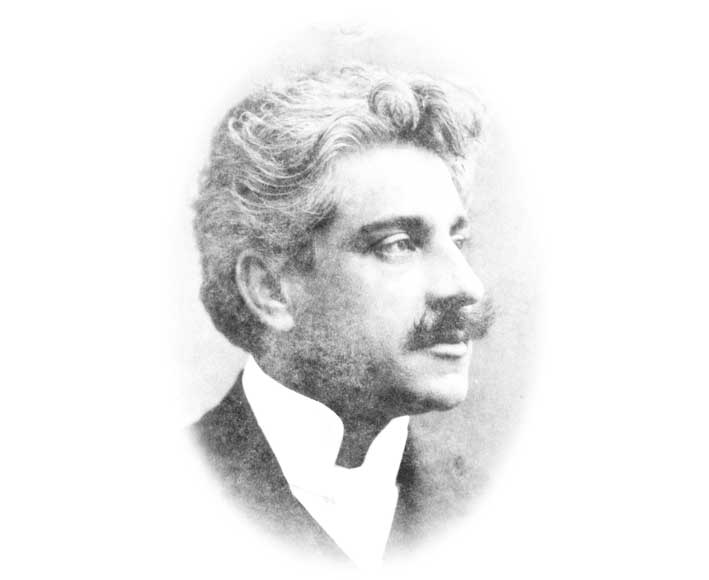 Em recorte redondo, nota-se o rosto de um senhor, de perfil. O homem tem cabelos grisalhos e ondulados, além de bigode; usa terno e gravata escuros e camisa clara.