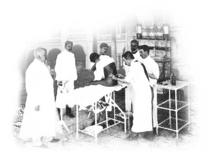 Foto de homens com longas vestimentas brancas ao redor de maca de hospital. Um deles examina uma pessoa que está sentada sobre o móvel.