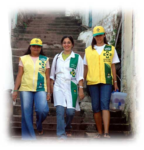 Três mulheres uniformizadas com jaleco e coletes do Programa Saúde da Família são o destaque desta imagem. As profissionais da saúde descem, lado a lado, uma longa escadaria. As três olham para a câmera e sorriem discretamente.