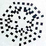 Nesta foto, observa-se uma mandala feita de pequenos quadrados pretos sobre fundo branco. Os quadriláteros foram posicionados lado a lado, de tal maneira que originaram formas circulares, umas dentro das outras, além de raios.