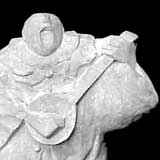 A forma de uma pessoa é reconhecida na escultura que compõe esta foto colorida. Numa pedra branca identifica-se os contornos de uma pessoa baixa e larga tocando banjo.