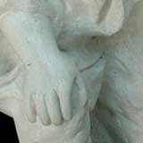 Uma peça em esculpida em pedra branca aparece nesta foto. Identifica-se um homem com um dos joelhos no chão e o outro flexionado, onde se apoia uma das mãos; a outra segura um objeto arredondado. Nota-se também traços do rosto dele, como olhos bem abertos, além de barba longa e bigode.