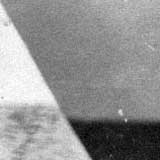 Nesta foto em preto e branco, identifica-se um objeto triangular à esquerda em frente a uma parede.