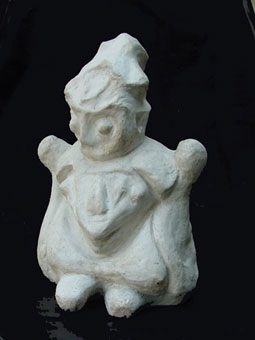 A fotografia revela escultura em material branco sobre fundo preto. A figura é um tanto disforme, contudo, identificam-se partes como cabeça, mãos e pés, remetendo à representação de uma forma humana.