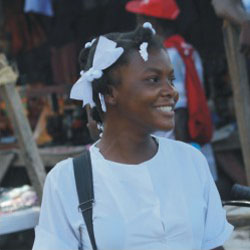 Aproximada e de perfil, uma adolescente negra estampa um largo sorriso, enquanto olha para a direita nesta foto. Notam-se laços de fita branca no cabelos, combinando com a blusa da mesma cor. Ao fundo, em desfoque, percebe-se uma espécie de feira, com bancas improvisadas repletas de objetos expostos. 