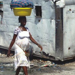 Esta foto mostra mulher gestante, jovem e negra carregando, sobre a cabeça, bacia com um avultado embrulho. Nota-se um contêiner ao fundo e resíduos de lixo espalhados pelo chão por onde ela caminha.