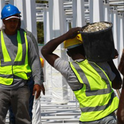 Nesta foto em close, funcionários de uma obra carregam baldes nos ombros. Usam capacetes azuis ou amarelos e coletes chamativos, também amarelos. Nota-se que deslocam-se no sentido do fundo da imagem, andando por entre as vigas de sustentação do prédio. À extrema esquerda, vê-se um homem em frente a uma máquina de misturar cimento. 