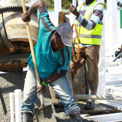 Dois homens negros trabalhando em uma obra são o destaque desta foto. Um deles está com as botas submersas na massa de cimento que maneja com o auxílio de uma pá. O outro, um pouco mais atrás, usa capacete, máscara e colete e está próximo a uma máquina de misturar cimento. Ao fundo, notam-se mais funcionários entre as vigas de sustentação do edifício.