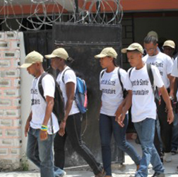 Nesta foto em close, vê-se em destaque um homem negro trabalhando em canteiro de obra. Ele está no centro da imagem, usando capacete e colete amarelos e manejando ferramenta para lidar com cimento. Atrás dele, entre as vigas de sustentação do prédio, outros trabalhadores são avistados.