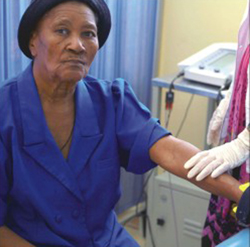 Uma senhora negra está sendo atendida por profissional de saúde branca nesta foto. A idosa usa blusa azul e chapéu preto. Sentada à esquerda, olha fixa e seriamente para a câmera, enquanto seu braço esquerdo fica estendido para frente. A mulher que a atende usa luva e jaleco brancos e tem um aparelho instalado na mão da paciente. 