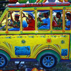 Esta foto mostra um ônibus amarelo de rodas azuis ocupado por passageiros e motorista negros. É uma peça artesanal decorativa focalizada em close. Na parte de cima lateral do carro, há a palavra “Delmas”.