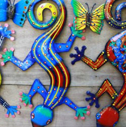 Nesta foto observam-se três salamandras artesanais e decorativas expostas sobre um fundo de madeira. Estão com a cabeça voltada para o chão. A primeira é azul e amarela com bolas brancas. A segunda é azul com listras amarelas e bolas brancas e vermelhas. Já a terceira, tem o corpo vermelho com bolas amarelas e patas azuis. Entre uma e outra peça, veem-se objetos menores igualmente coloridos, que representam borboletas.