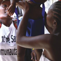 Encostadas a uma parede colorida, três jovens negras interagem. A foto mostra que a pintura atrás delas retrata uma garota negra que, talvez pelo ângulo da imagem, acaba confundindo-se com o trio. Na parte de cima da parede há a palavra “Kindergarten” 