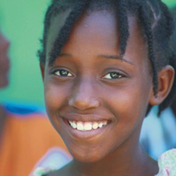 Nesta foto, o rosto de uma menina negra é o destaque. Ela está de frente para  câmera, esbanjando um sorriso aparentemente sincero. Os cabelos curtos e escuros são diferenciados pelo penteado rastafári.