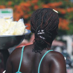 Em close, uma mulher negra está de costas para a câmera, equilibrando uma bacia sobre a palma da mão esquerda. Ela usa penteado rastafári e blusa azul de alça. Dentro da bacia, há pequenos sacos transparentes com alimentos. O fundo da imagem é totalmente desfocado.