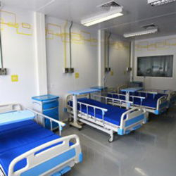 Nesta foto, pelo menos três camas de hospital e três criados mudos estão visíveis. Observa-se que ocupam parte de um ambiente grande, iluminado, com piso, paredes e teto brancos.