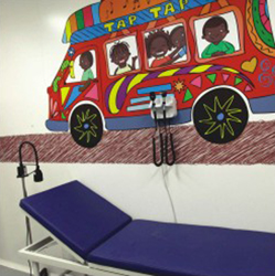 Esta foto apresenta o interior de um consultório. A parede ao fundo é decorada com o desenho de um ônibus coloridíssimo, dentro do qual estão passageiros, todos negros. Em cima do veículo notam-se cestas de frutas e as palavras “tap tap”. Logo abaixo da arte, vê-se maca azul e cadeira preta. À extrema direita da imagem, percebe-se um homem negro caminhando, pois a porta do consultório está aberta. O transeunte passa em frente a um grande painel de fotos criado na parede externa à sala.