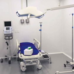 Uma sala com equipamentos hospitalares é vista nesta foto. À esquerda vê-se pia e compartimentos de sabonete e papel. Ao lado, máquina que mostra batimentos cardíacos e outras informações. Em seguida, observa-se maca e itens como suporte móvel para soro.