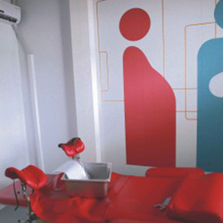 Nesta foto, identifica-se maca ginecológica em tecido vermelho. Na parede ao fundo, há uma pintura decorativa de bonecos representando gestante e médico. À direita, no canto, vê-se também uma cadeira preta. 