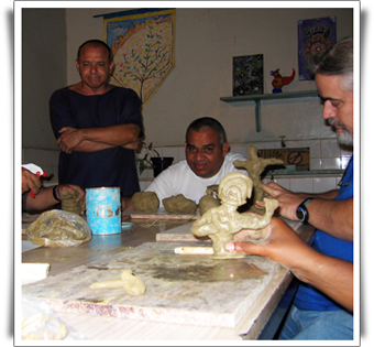 Nesta fotografia em cores três pessoas estão sentadas a uma mesa, trabalhando em esculturas pequenas em argila. Um homem está em pé, com os braços cruzados, observando o trabalho. Na sala, ao fundo, existe uma prateleira com algumas peças de materiais diversos. O homem que está sentado na cabeceira olha para a câmera e sorri.