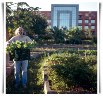 Fotografia em cores de um homem em uma horta. Ele sorri, enquanto segura um cesto plástico preto cheio de hortaliças. O homem tem cabelos cacheados até a altura dos ombros, barba e bigode grisalhos. Ao fundo, algumas árvores oferecem sombra à paisagem da horta.