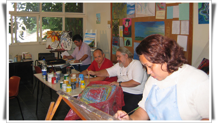 A fotografia mostra quatro pessoas em um ateliê de pintura. Em primeiro plano, uma mulher de cabelos castanhos, curtos e ondulados pinta em uma tela, que está apoiada sobre um cavalete. Ao fundo, três homens estão sentados a uma mesa onde há diversos potes de tinta, de cores variadas. Um deles está pintando um tecido, enquanto o homem ao lado o observa.