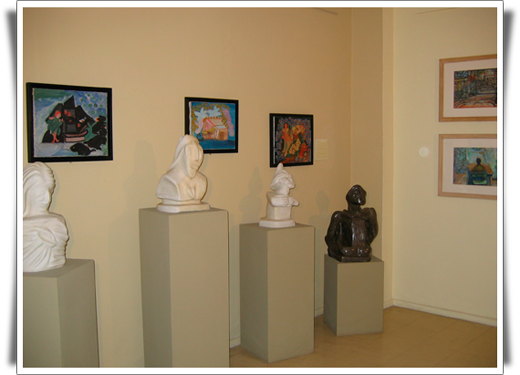 Esta foto em cores revela três bustos esculpidos em material branco e um em material escuro. Estão expostos juntamente a quadros bastante coloridos e afixados na parede.