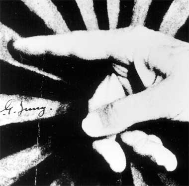 A fotografia, em preto e branco, apresenta o close da mão de Carl Jung apontando mandala pintada em uma tela. A mão, com o dedo indicador encostado no quadro, ocupa praticamente toda a área da imagem. Em um dos raios da mandala está a assinatura de Jung.