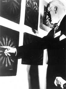 A fotografia está em preto e branco e mostra Carl Jung. Ele está de perfil, com a mão esquerda apoiada em uma bengala, enquanto aponta, com a mão direita, para mandala pintada em um dos quadros na parede. O quadro em destaque tem desenho de cor clara em fundo escuro e, além dele há mais quatro quadros afixados na parede.