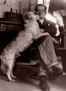 Fotografia em preto e branco de um senhor sentado em uma cadeira e um cachorro com as patas dianteiras sobre suas pernas. O homem está de frente para a câmera, fitando-a. Sua perna direita está cruzada sobre a esquerda, e as mãos estão sobre as patas do cachorro. O senhor, de cabelos curtos e escuros, tem a feição séria e veste blazer e sapatos escuros, camisa e calças claras. O cachorro aparece de perfil, é grande e tem pelos claros. Ao fundo, um objeto sobre uma bancada. Fotografia em preto e branco de um senhor sentado em uma cadeira e um cachorro com as patas dianteiras sobre suas pernas. O homem está de frente para a câmera, fitando-a. Sua perna direita está cruzada sobre a esquerda, e as mãos estão sobre as patas do cachorro. O senhor, de cabelos curtos e escuros, tem a feição séria e veste blazer e sapatos escuros, camisa e calças claras. O cachorro aparece de perfil, é grande e tem pelos claros. Ao fundo, um objeto sobre uma bancada.