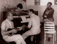 A imagem está em preto e branco e revela três homens tocando instrumentos musicais em uma sala. O homem que toca cavaquinho está mais à frente, sentado de perfil. Ele olha para a o pianista, à sua esquerda, que está sentado de costas. Em pé, à direita dele, há um homem tocando violão, que está com o pé direito sobre uma cadeira clara. Ele olha para a esquerda da imagem, onde, atrás do piano há uma pessoa em pé. O fundo da fotografia mostra, ao lado direito, uma parede clara bicolor com alguns quadros afixados e, do outro lado, onde está o piano, a pintura de uma paisagem. A imagem está em preto e branco e revela três homens tocando instrumentos musicais em uma sala. O homem que toca cavaquinho está mais à frente, sentado de perfil. Ele olha para a o pianista, à sua esquerda, que está sentado de costas. Em pé, à direita dele, há um homem tocando violão, que está com o pé direito sobre uma cadeira clara. Ele olha para a esquerda da imagem, onde, atrás do piano há uma pessoa em pé. O fundo da fotografia mostra, ao lado direito, uma parede clara bicolor com alguns quadros afixados e, do outro lado, onde está o piano, a pintura de uma paisagem.
