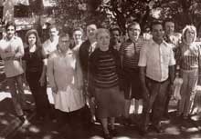A fotografia está em preto e branco e mostra a doutora Nise da Silveira junto a um grupo de pessoas. Todos estão em pé, sob a sombra de algumas árvores. Ela encontra-se no centro, tem os cabelos grisalhos presos, usa óculos redondos, blusa escura listrada na horizontal, saia e sapatos também escuros. Ao fundo, árvores e uma construção branca. A fotografia está em preto e branco e mostra a doutora Nise da Silveira junto a um grupo de pessoas. Todos estão em pé, sob a sombra de algumas árvores. Ela encontra-se no centro, tem os cabelos grisalhos presos, usa óculos redondos, blusa escura listrada na horizontal, saia e sapatos também escuros. Ao fundo, árvores e uma construção branca.