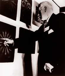 A fotografia está em preto e branco e mostra Carl Jung, de perfil, com a mão esquerda apoiada em uma bengala e, com a direita, aponta para mandala pintada em um dos quadros na parede. O quadro em destaque tem fundo escuro e o desenho é de cor clara. Há mais quatro quadros afixados na parede. A fotografia está em preto e branco e mostra Carl Jung, de perfil, com a mão esquerda apoiada em uma bengala e, com a direita, aponta para mandala pintada em um dos quadros na parede. O quadro em destaque tem fundo escuro e o desenho é de cor clara. Há mais quatro quadros afixados na parede.