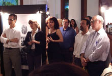 A foto, em cores, apresenta um grupo de pessoas em pé, olhando para o lado esquerdo da imagem. Ao fundo, há um painel e mais pessoas em pé.