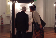 Fotografia colorida de duas pessoas observando um painel da exposição. No centro da foto, um homem de costas e uma mulher de perfil olham para um painel que contém um longo texto. A mão direita da mulher está em movimento, apontando para o painel. Na imagem também se percebem o piso de madeira, as paredes brancas e um extintor de incêndio na lateral esquerda, sobre o chão.