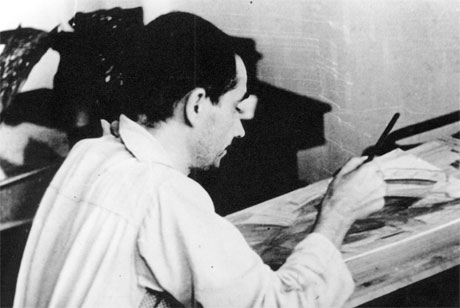 A fotografia em preto e branco mostra um homem de meia-idade sentado, de perfil. Na mão direita, segura um pincel, que está em contato com uma tela. Ele tem cabelos curtos e escuros e veste camisa de manga comprida clara. Ao fundo, há uma mesa com alguns objetos.