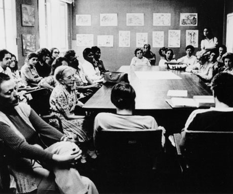 A fotografia, em preto e branco, mostra um grupo de mais de vinte pessoas reunidas em uma sala. Estão ao redor de uma grande mesa retangular de madeira, com a presença da doutora Nise da Silveira, já idosa. À esquerda, há duas janelas e dois quadros. Ao fundo, uma parede está tomada por doze quadros.