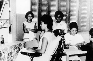 Nesta fotografia, em preto e branco, há cinco mulheres. Em primeiro plano, uma delas está sentada, de perfil, e olha para frente. Ela tem cabelos curtos e veste roupas claras. Ao fundo, há duas mulheres, também de cabelos curtos e roupas claras, ambas em pé, com a feição séria e os braços cruzados sobre o peito. No canto direito da imagem, uma mulher faz o trabalho de manicure em outra.