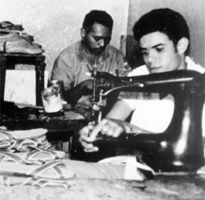 A fotografia em preto e branco mostra dois homens exercendo o ofício de sapateiro. À direita, em primeiro plano, há uma máquina de costura preta, na qual trabalha um homem de cabelos escuros e camisa clara. No canto inferior esquerdo, existem algumas sandálias empilhadas. Ao fundo, um homem também está costurando e, ao lado dele, há diversas formas de sapatos em madeira sobre uma prateleira.