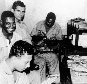 Fotografia, em preto e branco, mostra quatro sapateiros jovens trabalhando. Eles usam uniforme hospitalar. O homem que está em primeiro plano tem expressão séria, e olha para a sandália que traz nas mãos. O segundo, olha para a câmera, com um largo sorriso no rosto. O homem ao lado deste, também tem nas mãos uma sandália, e olha para a câmera, porém está sério. O último está martelando uma sandália. À direita, há uma prateleira, onde estão várias sandálias e formas de sapato em madeira.