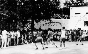 A fotografia em preto e branco mostra homens jogando uma partida de futebol. Uma torcida numerosa assiste ao jogo, que acontece em uma quadra enfeitada com bandeirolas juninas. A copa de uma árvore grande oferece sombra à boa parte das pessoas. Os jogadores vestem uniformes compostos de bermudas, camisetas e meias compridas com listras horizontais. Um jogador, posicionado no meio do campo, veste meias curtas. Ao fundo, há uma construção branca com um muro longo e alto, e algumas janelas.