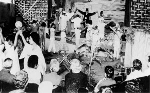 Nesta fotografia em preto e branco, homens e mulheres estão dançando em um palco e vestem roupas claras. No centro deste tablado, há uma pessoa de fraque e cartola escuros. Todos estão de mãos dadas, com os braços erguidos. Pessoas sentadas de costas para a câmera estão assistindo à apresentação.