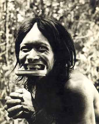 Nesta foto em preto e branco, um pessoa indígena sorri para a câmera. Notam-se a falta de dentes e o alargamento do lábio inferior com o uso de objeto em forma de disco.