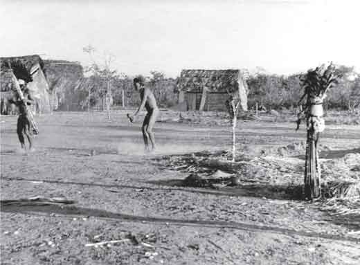 Fotografia apresenta indígenas em aldeia onde se vê espécie de totem.