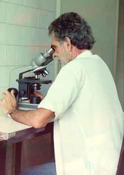 A foto traz um homem sentado à bancada observando através de um microscópio.