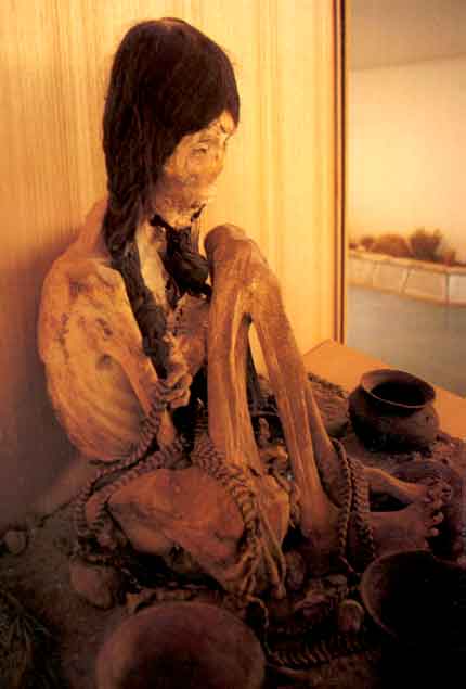 A foto revela um esqueleto ainda vestido, com cabelos longos e trançados. A ossada é de uma pessoa que estava sentada no chão, próxima a vasilhas.