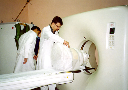 Foto em cores de dois profissionais da saúde realizando exame de tomografia computadorizada em um volume coberto com tecido branco. 