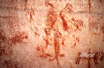 Imagem revela desenhos rupestres de pessoas com braços estendidos e elevados. Notam-se ainda outros indivíduos ao redor de um volume, tocando-o, também com os braços retesados.