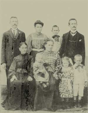  Foto em preto e branco de adultos e crianças. Lado a lado e posando seriamente para a câmera, alguns estão em pé e outros sentados. Ao todo, há nove pessoas: quatro adultos, um adolescente e quatro crianças.  A fotografia é bastante antiga, o que explica roupas, penteados e posturas do século passado. 